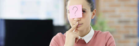 Pensive young woman sitting at table with sticker with question mark on her foreheadの写真素材