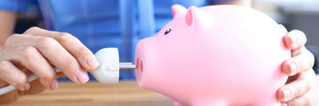 Woman is holding electrical plug and and pink piggy bank closeup