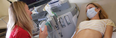 Woman in protective medical mask lying on couch in front of doctor with ultrasound diagnosticsの写真素材