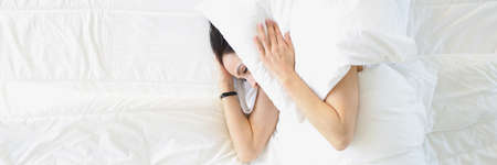 Young woman lying in white bed and covering her ears with pillow top view
