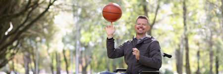 Smiling disabled man twirls basketball ball on his finger while sitting in wheelchair in park
