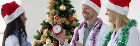 Group of doctors in santa claus hats standing near christmas tree and holding red alarm clock and...