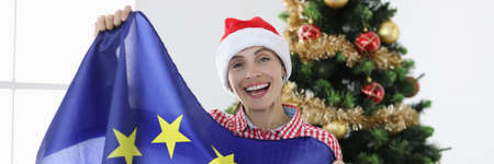 Joyful woman in santa claus hat holds european union flag against background of christmas treeの写真素材