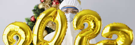 Doctor in protective suit, mask and glasses holds numbers 2022 against background of New Year treeの写真素材