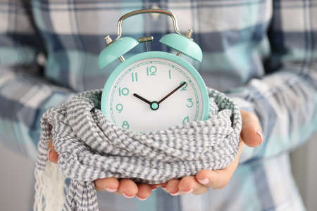 Lady hold green alarm clock, changing time from summer to winter, clock wrapped in knitted scarf