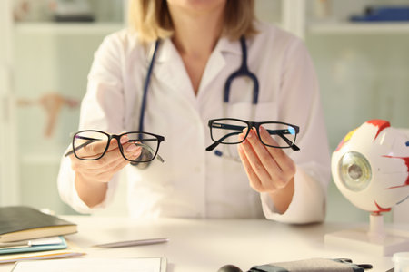 Ophthalmologist holds different eyeglasses in her hands while sitting at desk in medical clinic.の写真素材