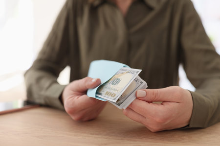 Woman sits at table and holds envelope with stack of dollar bills close-up.