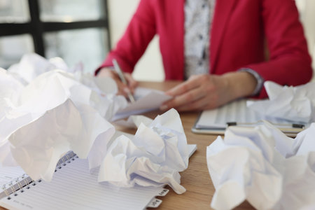 Woman sits at wooden table near scattered crumpled papersの写真素材