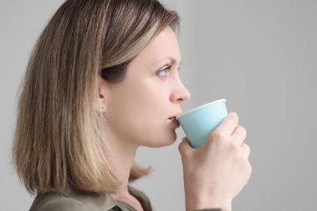 Blonde woman drinks coffee from blue paper cup in morningの写真素材