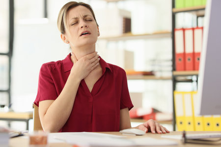 Woman touches painful throat sitting at desk in officeの写真素材