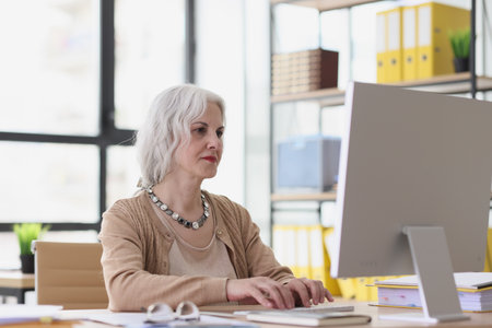Woman with grey hair looks at screen of computer monitorの写真素材