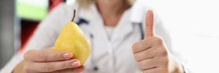 Nutritionist woman showing yellow pears and thumbs up gesture while sitting at medical clinic table.の写真素材