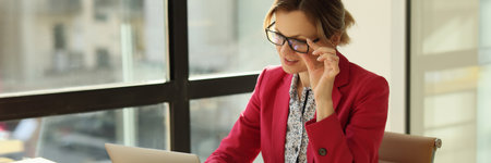 Female manager sits at her workplace in office, touches her glasses and looks at laptop screen.の写真素材