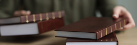 Woman puts thick books with brown cover on wooden tableの写真素材
