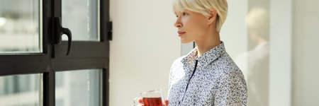 Thoughtful woman with tea cup looks out of window in officeの写真素材