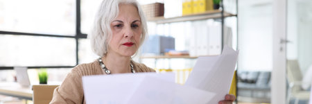 Serious grey haired woman studies reports sitting at deskの写真素材