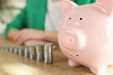 Piggy bank and stacks of coins on wooden table, business person in background.の写真素材