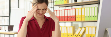 Suffering woman massages temples with hands sitting at deskの写真素材