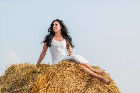 Beautiful brunette woman sitting on hay bale in warm summer dayの写真素材