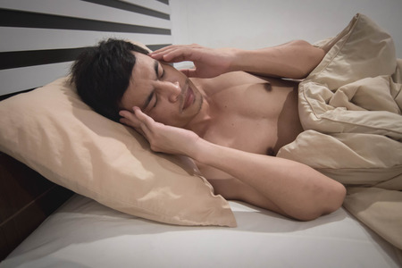 Young Asian Caucasian man on bed having headache / insomnia / migraine / stress / broken.の写真素材