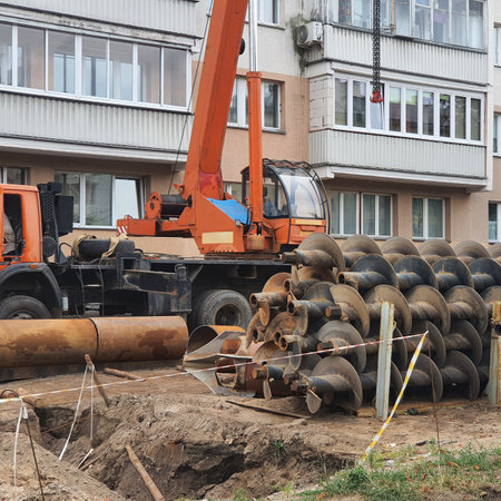 Orange crane truck lifting screw piles near a residential building, preparing for foundation installation. Heavy machinery working diligently at the construction site for urban developmentの写真素材