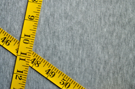 The measuring tape of yellow color with numerical indicators in the form of centimeters or inches lies on a gray knitted fabric. Background concept for sewing clothes for specific body sizesの写真素材