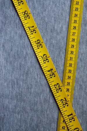 The measuring tape of yellow color with numerical indicators in the form of centimeters or inches lies on a gray knitted fabric. Background image about sewing sportswear for people, who slimmingの写真素材
