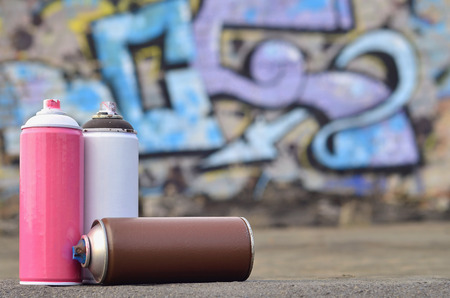 A still-life of several aerosol paint spray cans lies on a roofing covering on the background of a brick wall, on which is painted a bright and colorful graffiti drawing. Items related to street artの写真素材