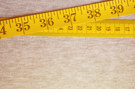 The measuring tape of yellow color with numerical indicators in the form of centimeters or inches lies on a gray knitted fabric. Background concept for sewing clothes for specific body sizesの写真素材