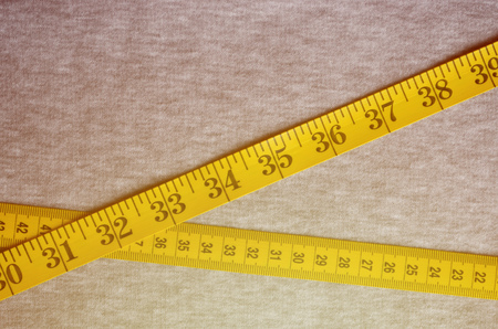 Yellow measuring tape with numerical indicators in the form of centimeters or inches lies on a gray knitted fabric. Concept industry associated with sewing sportswear for specific body sizesの写真素材