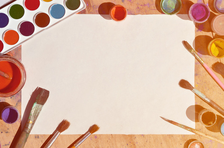 Background image showing interest in watercolor painting and art. A blank sheet of paper, surrounded by brushes, cans with watercolor paint and gouache, which lie on an old and stained wooden tableの写真素材
