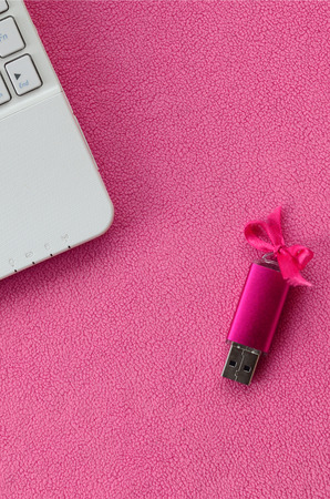 Brilliant pink usb flash memory card with a pink bow lies on a blanket of soft and furry light pink fleece fabric beside to a white laptop. Classic female gift design for a memory cardの写真素材