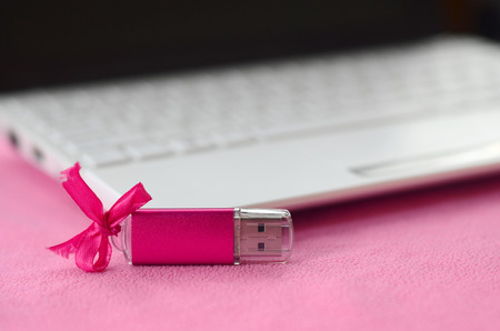 Brilliant pink usb flash memory card with a pink bow lies on a blanket of soft and furry light pink fleece fabric beside to a white laptop. Classic female gift design for a memory cardの写真素材