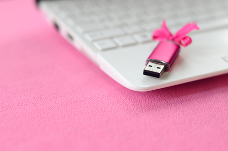Brilliant pink usb flash memory card with a pink bow lies on a blanket of soft and furry light pink fleece fabric beside to a white laptop. Classic female gift design for a memory cardの写真素材