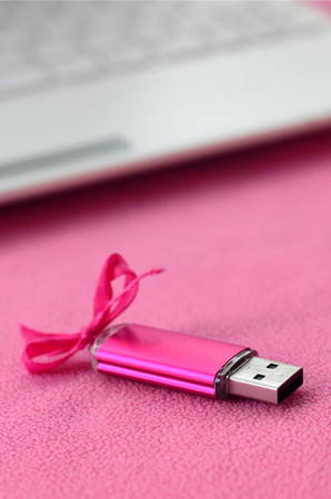 Brilliant pink usb flash memory card with a pink bow lies on a blanket of soft and furry light pink fleece fabric beside to a white laptop. Classic female gift design for a memory cardの写真素材