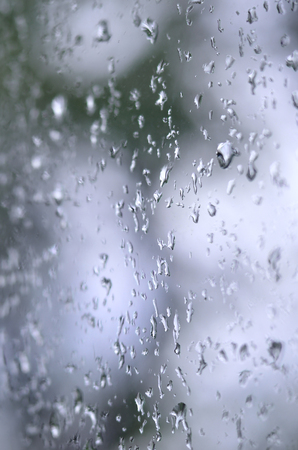 A photo of rain drops on the window glass with a blurred view of the blossoming green trees. Abstract image showing cloudy and rainy weather conditionsの写真素材