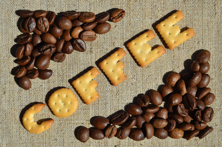 Background of a gray canvas made of old and coarse burlap, on which lies a certain amount of brown coffee beans and the word "coffee", composed of edible letters made of yellow salted crackerの写真素材
