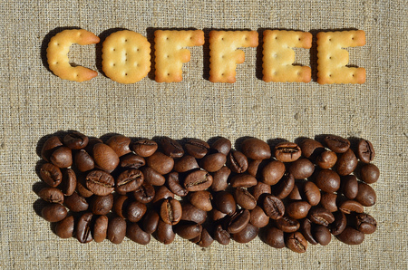 Background of a gray canvas made of old and coarse burlap, on which lies a certain amount of brown coffee beans and the word "coffee", composed of edible letters made of yellow salted crackerの写真素材