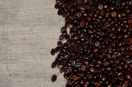 Texture of a gray canvas made of old and coarse burlap, on which lies a certain amount of brown coffee beans. Top view of sackcloth of light gray color under sunlight with pile of coffee beansの写真素材