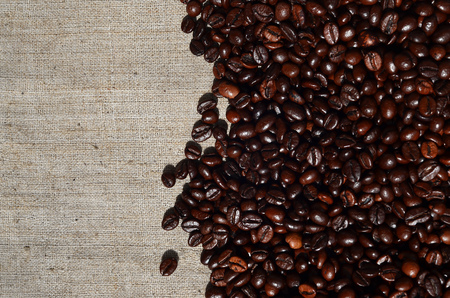 Texture of a gray canvas made of old and coarse burlap, on which lies a certain amount of brown coffee beans. Top view of sackcloth of light gray color under sunlight with pile of coffee beansの写真素材