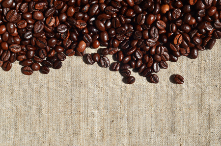 Texture of a gray canvas made of old and coarse burlap, on which lies a certain amount of brown coffee beans. Top view of sackcloth of light gray color under sunlight with pile of coffee beansの写真素材