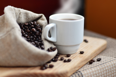 A full bag of brown coffee beans and a white cup of hot coffee lies on a wooden surface. Attributes related to the preparation of natural coffeeの写真素材