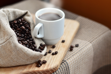 A full bag of brown coffee beans and a white cup of hot coffee lies on a wooden surface. Attributes related to the preparation of natural coffeeの写真素材