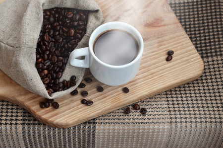 A full bag of brown coffee beans and a white cup of hot coffee lies on a wooden surface. Attributes related to the preparation of natural coffeeの写真素材