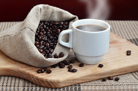 A still life with a bag full of brown coffee beans, which lies next to a small white cup of hot coffee on a wooden board behind a small low table covered with a checkered tableclothの写真素材