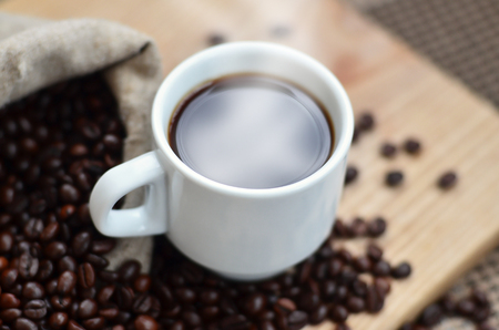 A still life with a bag full of brown coffee beans, which lies next to a small white cup of hot coffee on a wooden board behind a small low table covered with a checkered tableclothの写真素材