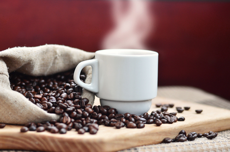 A still life with a bag full of brown coffee beans, which lies next to a small white cup of hot coffee on a wooden board behind a small low table covered with a checkered tableclothの写真素材