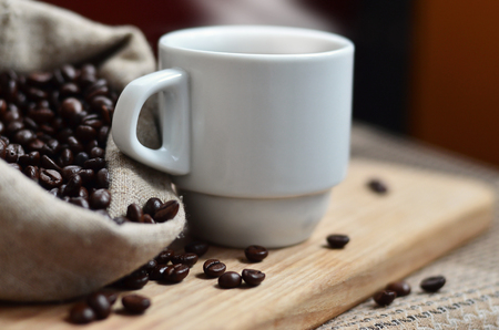 A full bag of brown coffee beans and a white cup of hot coffee lies on a wooden surface. Attributes related to the preparation of natural coffeeの写真素材