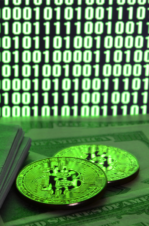 Two bitcoins lies on a pile of dollar bills on the background of a monitor depicting a binary code of bright green zeros and one units on a black background. Low key lightingの写真素材