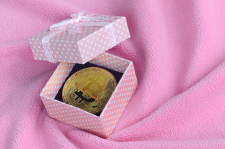 The golden bitcoin lies in a small pink gift box with a small bow on a blanket made of soft and fluffy light pink fleece fabric with a large number of relief foldsの写真素材